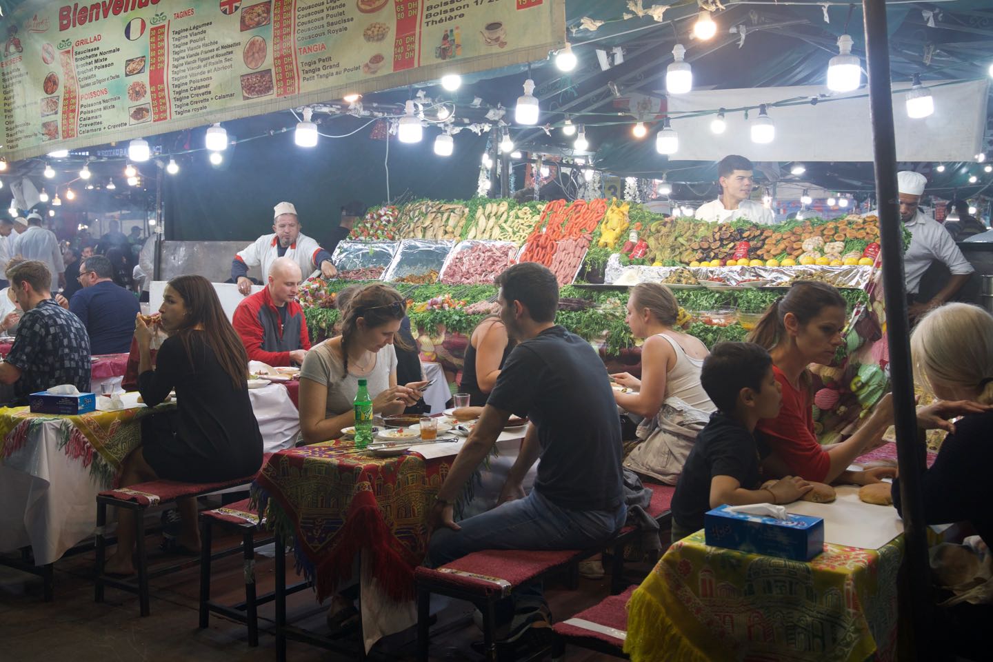 People seating on the tables of a food stall at Jemaa el-Fnaa at night.